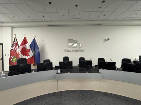 Town of Newmarket council chambers with a curved council desk, computer monitors and the Ontario, Canadian and Newmarket flags displayed beside the Newmarket logo.