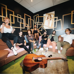 A group of seven people sitting on couches in a stylish room with a guitar on the table.