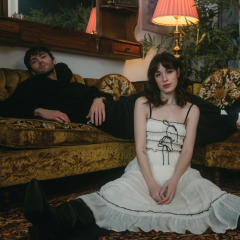Man reclining on retro sofa, woman seated on floor in white dress, cozy room lighting.