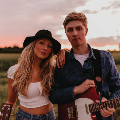 Two people with guitars in a field at sunset. One wears a hat.