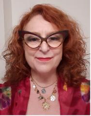 Woman with red hair, wearing large glasses and layered necklaces, smiling slightly.