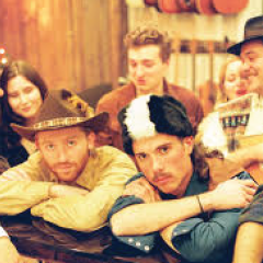 Group of people in cowboy attire, leaning on a piano with a rustic background.