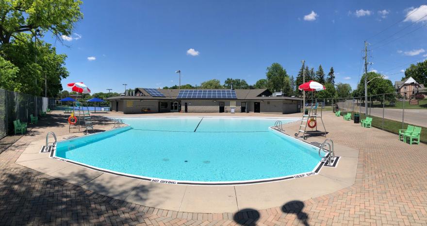Public swimming pool with blue water, sunny day, green benches, and red umbrellas.