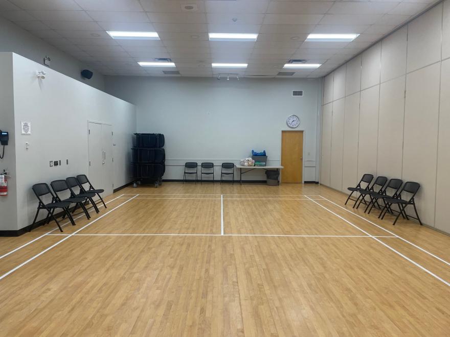 Empty room with wooden floor, folding chairs along walls, and a table at the back.