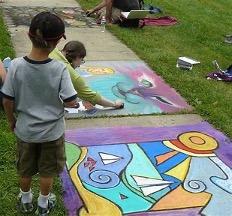 Child watches colorful chalk art on sidewalk grass.