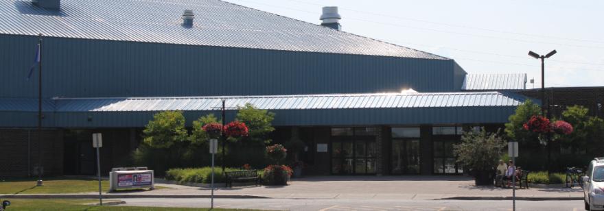 Large building with a metal roof, entrance flanked by flowering plants and streetlights.